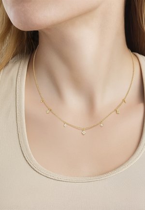 Sottile collana a catena d'oro con piccoli ciondoli a quadrifoglio pendenti indossata al collo di una donna sopra una maglietta beige.