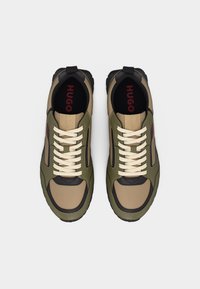 Ζευγάρι μπεζ και χακί αθλητικά παπούτσια με κορδόνια, με μαύρες λεπτομέρειες και κόκκινη επιγραφή "HUGO" στις μαύρες εσωτερικές σόλες.