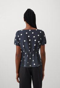 Blouse bleue à pois avec des manches courtes bouffantes, taille élastique et décolleté carré, ornée de motifs circulaires blancs sur un tissu doux.