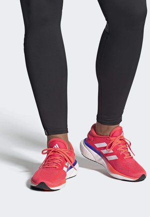 Zapatillas deportivas de un rosa brillante con rayas blancas, parte superior de malla texturada, suela acolchada y acentos contrastantes en púrpura y negro.