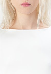 Hvit, glatt plagg med bred utringning som sitter tett til overkroppen. Myk tekstur; hår delvis skjuler skuldrene.