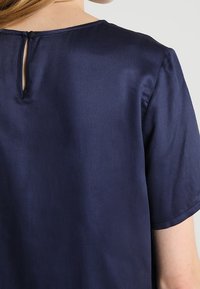 Bakvy av person som bär en mörkblå satinblus med kort ärm och en liten knappad nyckelhålöppning vid halsringningen.