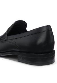 Zapato de vestir negro de cuero tipo mocasín con tacón bajo, costuras visibles y logotipo "BOSS" en relieve en el lateral de la suela.