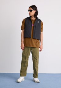 Vesta neagră fără mâneci cu fermoare, purtată cu un tricou maro și pantaloni verde măsliniu. Adidașii albi completează ținuta.