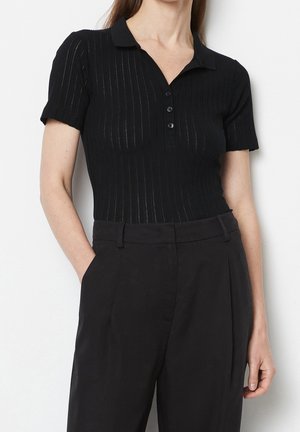 Vrouw in een zwarte polo met korte mouwen en ribbelstructuur met drie knopen, gestoken in hoge zwarte broek, met één hand in de zak.