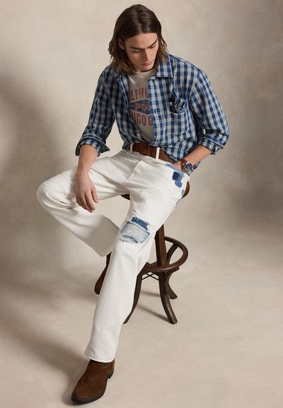 Νέος άνδρας με μαλλιά μέχρι τους ώμους κάθεται σε ξύλινο σκαμπό φορώντας μπλε καρό πουκάμισο, μπλουζάκι με γραφικό σχέδιο, λευκό distressed τζιν, καφέ ζώνη και μπότες.