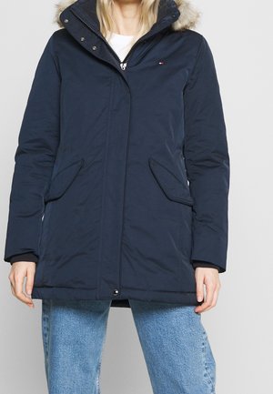 Femme portant une veste d'hiver bleu marine avec capuche bordée de fourrure, fermeture éclair devant, boutons pression et poches à rabat en biais, associée à un jean bleu clair.