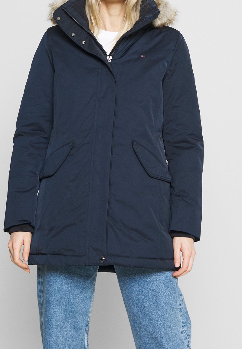 Femme portant une veste d'hiver bleu marine avec capuche bordée de fourrure, fermeture éclair devant, boutons pression et poches à rabat en biais, associée à un jean bleu clair.
