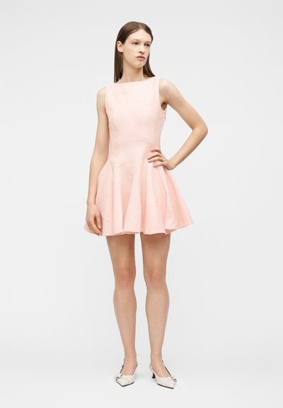 Γυναίκα που στέκεται με το χέρι στη μέση, φορώντας αμάνικο ροζ ανοιχτόχρωμο φόρεμα σε σχήμα Α με υφή και λευκά μυτερά τακούνια σε μονόχρωμο φόντο.