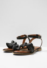 Czarne skórzane sandały z falbaniastymi akcentami, otwarte z przodu, z paskiem wokół kostki oraz płaską, brązową podeszwą. Charakteryzują się eleganckim i prostym wzornictwem.