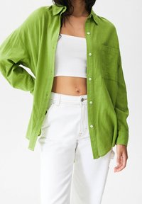 Chemise verte à manches longues en tissu léger, avec une coupe ample et une poche poitrine unique, associée à un pantalon blanc taille haute.