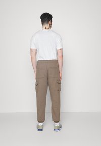 Brown cargo pants with elastic waistband and multiple side pockets, worn with a white T-shirt and grey sneakers. Back view.