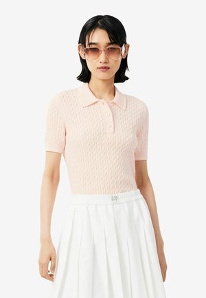Vrouw draagt een lichtroze gebreide polo met korte mouwen, witte geplooide rok en grote vierkante gradient zonnebril, staande tegen een effen achtergrond.