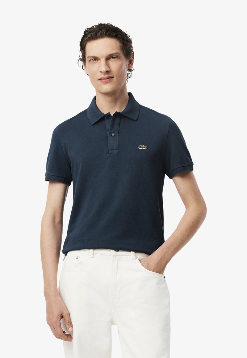 Camisa polo azul marino de algodón con cuello abotonado, mangas cortas y un pequeño logo de cocodrilo verde en el lado izquierdo del pecho. Lleva puesta con jeans blancos.