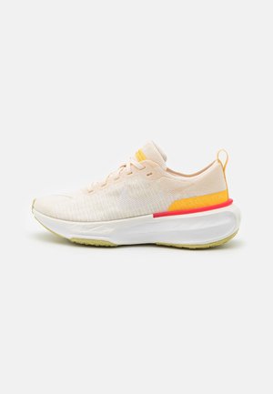 Light beige running shoe with a knit upper, yellow accents, white midsole, and a vibrant red detail on the heel. Textured surface.
