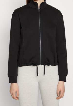 Black zip-up jacket with long sleeves and drawstring hem worn over light gray pants, front view from neck to hips.