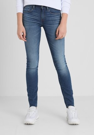 Person trägt Slim-Fit blaue Jeans und weiße Sneaker, steht mit entspannt an den Seiten hängenden Händen vor neutralem Hintergrund.