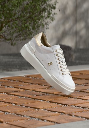 Pantof sport alb din piele cu accente aurii, ridicat pe o suprafață din dale de lemn cu pietricele mici între ele, cu un fundal neclar alcătuit dintr-un perete gri și copaci.