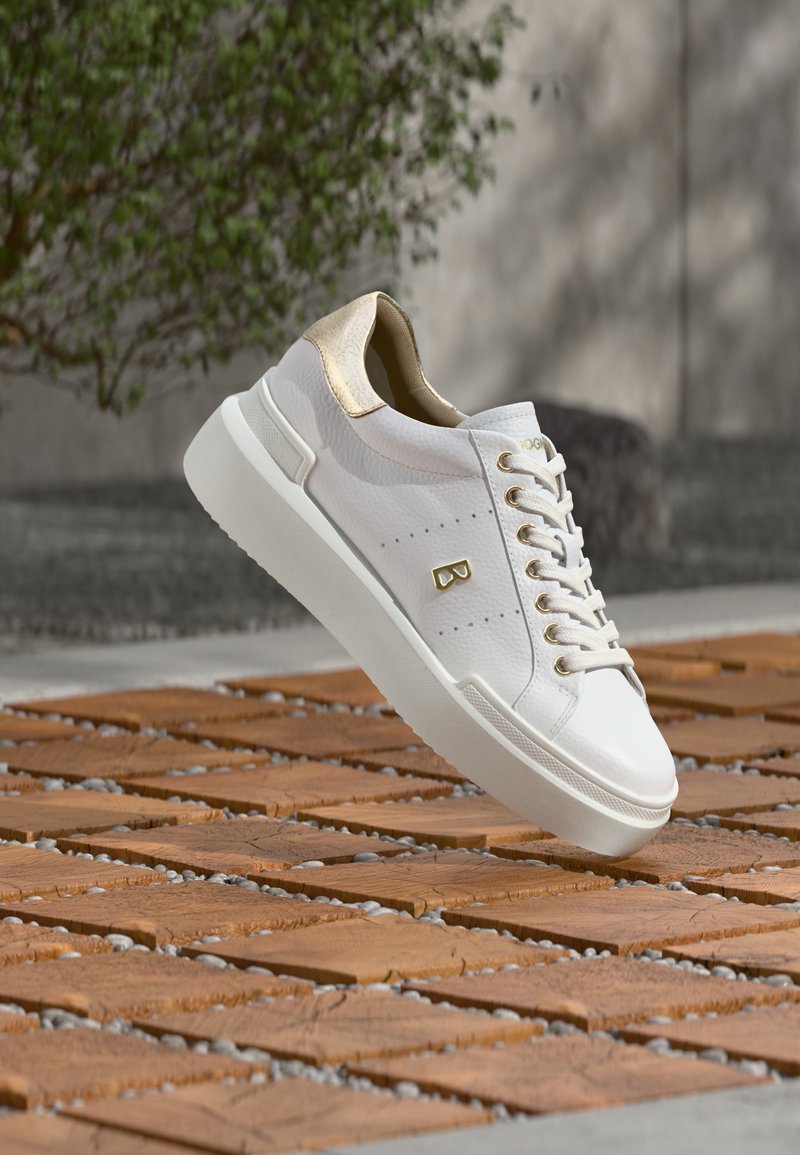 Baskets en cuir blanc avec des accents dorés, surélevées sur une surface de carreaux en bois avec de petits cailloux entre les carreaux, avec un mur gris flou et un arrière-plan d'arbres.