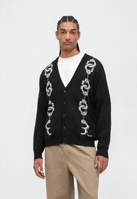 Un jeune homme avec des cheveux tressés porte un cardigan noir avec un motif de dés et de chaînes, une chemise blanche, et un pantalon beige, se tenant devant un fond uni.