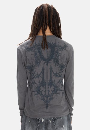 Personne avec de longues dreadlocks portant un t-shirt gris à manches longues arborant un design sombre, complexe, symétrique de ronces et d'épines dans le dos.