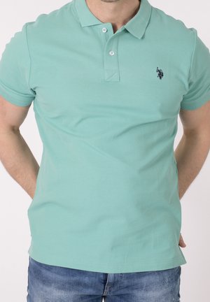 Mand iført en mintgrøn kortærmet poloshirt med mørkt logo på brystet og blå jeans, hænderne bag ryggen, neutral baggrund.
