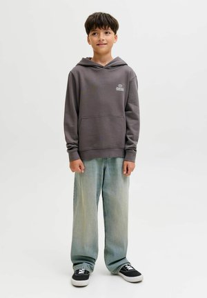 Ragazzo in piedi che indossa un felpa grigia con cappuccio, jeans blu chiaro larghi e sneakers nere su uno sfondo bianco semplice.