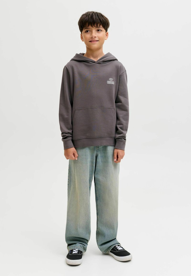 Ragazzo in piedi che indossa un felpa grigia con cappuccio, jeans blu chiaro larghi e sneakers nere su uno sfondo bianco semplice.