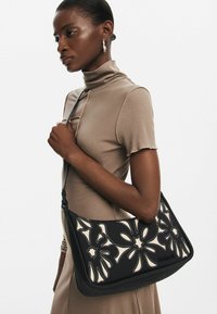 Borsa a spalla nera con design floreale intagliato al laser, dettagli color crema e texture liscia. Tracolla regolabile e chiusura con zip.