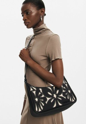 Sacoche noire avec un design de fleurs découpées au laser, accents crème et texture lisse. Bandoulière réglable et fermeture éclair.