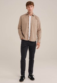 Cămașă bej cu nasturi, având două buzunare pe piept, stilizată peste un tricou alb, asociată cu pantaloni negri și adidași negri. Fundal simplu.