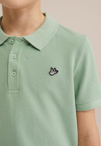 Svetlozeleno polo majica z teksturirano tkanino, tri gumbna zanka in črnim vezenim listnatim logotipom na prsih.