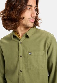 Camisa verde de botones en un tejido texturizado con un bolsillo de parche y un pequeño logo. Presenta un cuello clásico y botones marrones.
