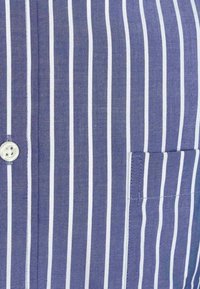 Blauw-wit gestreepte shirt met een enkele knoop bij de kraag en een patchzak aan de linkerkant. De stof lijkt gestructureerd.