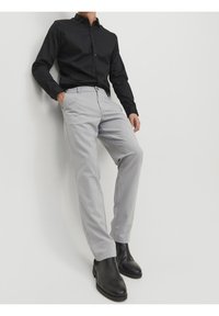 Camicia nera a maniche lunghe con bottoni, pantaloni grigi chiari con un motivo sottile e stivali neri alla caviglia; l'outfit è elegante e strutturato.