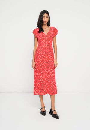 Rød blomstret midi-kjole med flæseærmer, V-udskæring og elastisk talje. Kombineret med sorte sko med rem. Enkelt hvidt baggrund.