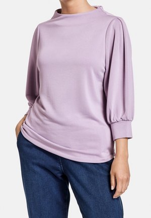 Kvinde iført en lyse lilla bluse med lange ærmer og puffede ærmer og mørkeblå jeans, står med den ene hånd i lommen.