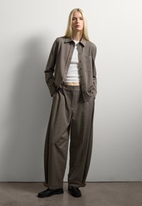 Giovane donna in piedi contro un muro bianco, con indosso un completo marrone morbido, camicia sbottonata, top corto bianco e mocassini neri, con le mani in tasca.