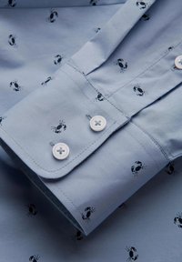 Camisa de color azul claro, hecha de algodón, que presenta un patrón de pequeñas ilustraciones de cangrejos negros. Incluye botones blancos en los puños.