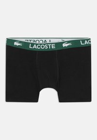 Sorte bomuldsboxershorts med en grøn linning, der har "LACOSTE" tekst og logo. Glat tekstur, midt-lår længde og tætsiddende pasform.