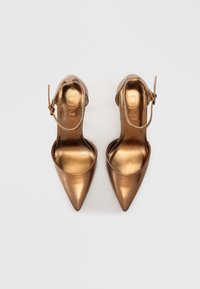 Brončane metalik cipele s visokom petom, s šiljastim vrhom i remenom oko gležnja. Glatka tekstura, moderan dizajn, s utisnutim logotipom na ulošcima.