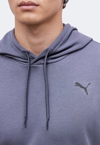 Gros plan d'une personne portant un sweat à capuche gris avec un logo Puma noir sur la poitrine et des cordons pendants.