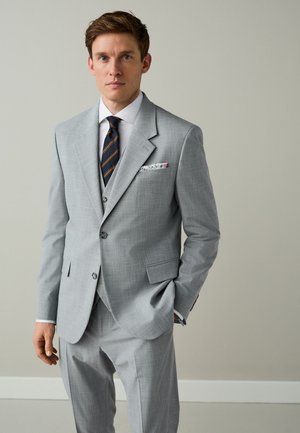 Hombre con traje gris claro con chaleco, camisa blanca, corbata oscura con rayas y un pañuelo floral en el bolsillo, de pie frente a un fondo neutro.