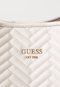 Sac à main en cuir beige matelassé avec couture en relief, logo doré "GUESS" et détails "EST 1981", texture lisse et bords doux.