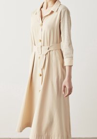 Abito beige con bottoni, maniche lunghe in rete testurizzata, vita delineata da una cintura e una gonna fluida, realizzato in tessuto leggero.