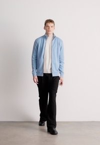 Ανοιχτό μπλε μπουφάν με φερμουάρ πάνω από λευκό ριγέ πουλόβερ, συνδυασμένο με μαύρο παντελόνι και μαύρα παπούτσια. Το μοντέλο περπατάει μπροστά σε ουδέτερο φόντο.