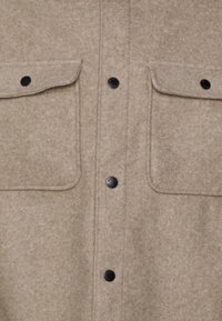 Tan wollen shirt met een knoopsluiting aan de voorkant, twee borstzakken en zwarte drukknoopsluitingen. De stof heeft een zachte, textuurachtige uitstraling.