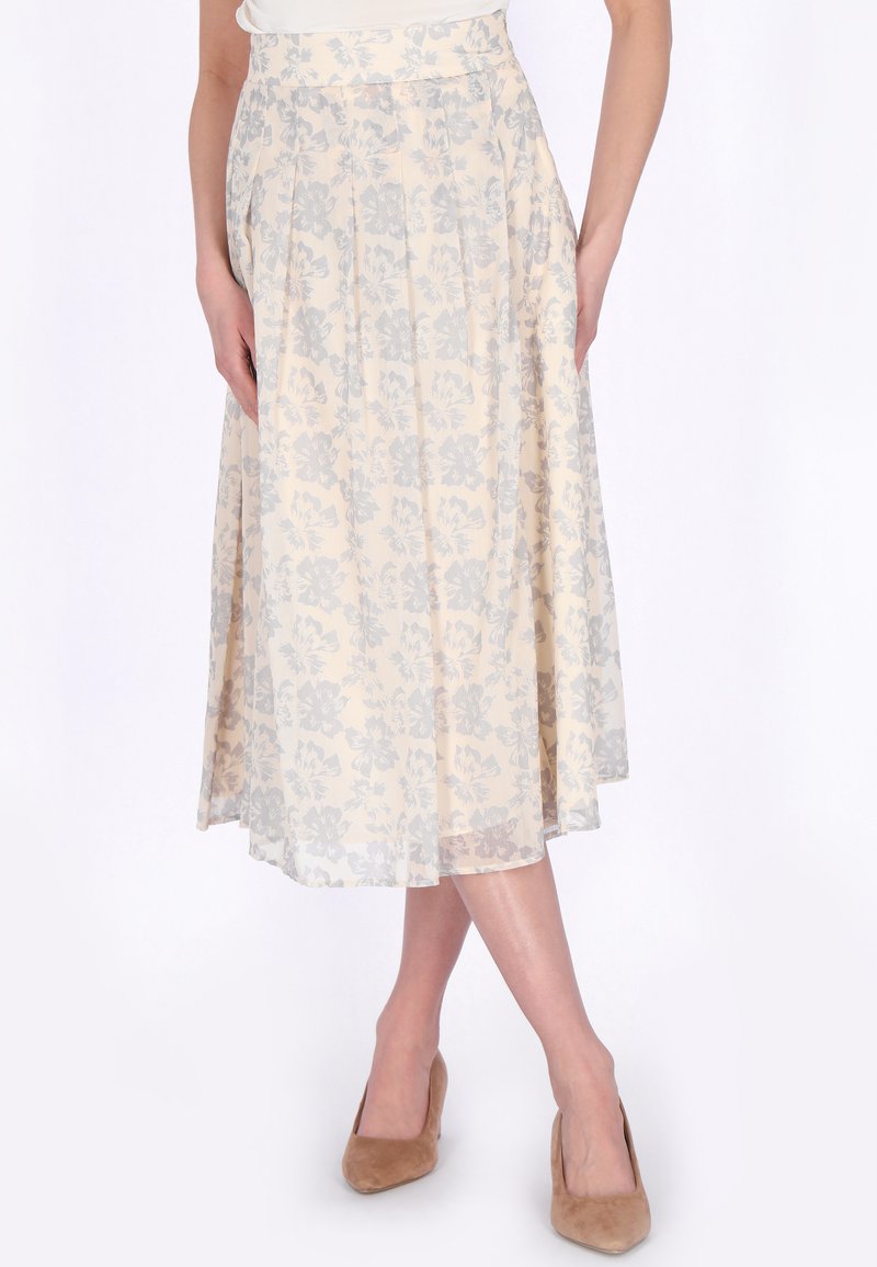 Hellbeiger Midirock mit grauem Blumenmuster, hoher Taille, Falten, getragen mit beigen Wildleder-Absätzen, gezeigt an einer Person mit gekreuzten Beinen.