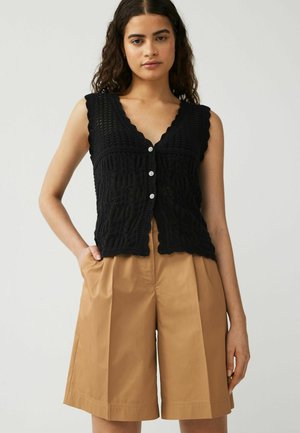 Zwarte, gebreide mouwloze top met een v-hals, golvende randen en drie decoratieve knopen. Gecombineerd met beige, hoog-gepaste shorts.