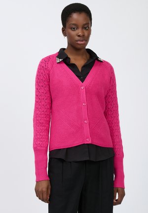Cárdigan de color rosa brillante con cuerpo texturizado, mangas con encaje y escote en V. Cuenta con cinco botones y puños acanalados. Lleva puesto sobre una camisa negra.